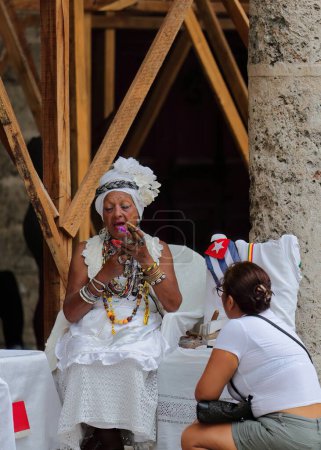 Foto de La Habana, Cuba-7 de octubre de 2019: Santera cubana con clavos espectaculares atiende a un cliente solicitando sus servicios de adivinación mientras fuma un gran cigarro bajo la galería de la Plaza Catedral lado este. - Imagen libre de derechos