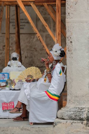 Foto de La Habana, Cuba-7 de octubre de 2019: Santera cubana con clavos espectaculares espera a que sus clientes soliciten sus servicios de adivinación y fuma un gran cigarro debajo de la Plaza Catedral. - Imagen libre de derechos