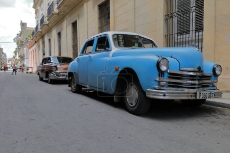 Foto de La Habana, Cuba-8 de octubre de 2019: American 4-door, hard top classic cars -blue Plymouth Special DeLuxe Sedan 1949, brown Chevrolet Bel Air Sedan 1957- stop on a Centro street while locals play baseball. - Imagen libre de derechos