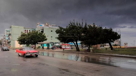 Foto de La Habana, Cuba-8 de octubre de 2019: El viejo auto clásico americano blanco-rojo almendro, tanque de yanqui Plymouth Savoy Sedán de 4 puertas 1958 conduce por la calle San Lázaro pasando por el Parque Antonio Maceo después de un fuerte aguacero. - Imagen libre de derechos
