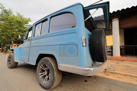 Foto de Vinales, Cuba-9 de octubre de 2019: Blue American classic car-almendron- Jeep Willys Station Wagon 4Wheel Drive desde 1955 con la puerta trasera superior abierta aparcada en una calle del centro de la ciudad al comienzo del atardecer. - Imagen libre de derechos
