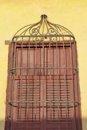 Trinidad, Kuba - 12. Oktober 2019: Braunbemalte Holzläden eines geschlossenen Fensters, geschützt durch ein starkes schmiedeeisernes Gitter, gelbe Fassade eines Kolonialhauses, Straße im Viertel Plaza Mayor.