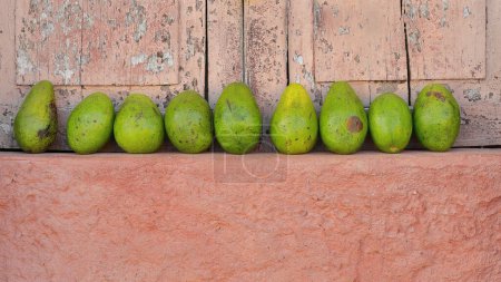 Green avocados, Catalina variety, lined on the salmon-orange colored ledge of a window with light pink, paint-chipped closed wooden shutters, for sale on the Plaza Mayor Square area. Trinidad-Cuba.