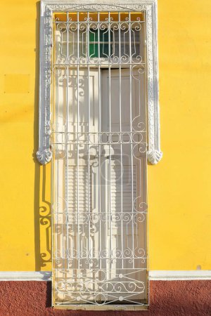 Trinidad, Cuba-October 12, 2019: White-painted wooden shutters, wrought iron grate and millwork frame of a closed tall window, yellow facade of a colonial house, street in the Plaza Mayor Square area.