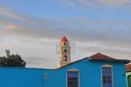 Trinidad, Cuba-12 de octubre de 2019: La torre de la antigua Iglesia de San Francisco de Asis sobresale sobre el muro azul cerúleo de una casa colonial de un piso en la Plaza Mayor, lado oeste.