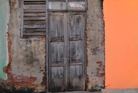Trinidad, Cuba-October 12, 2019: Dilapidated wood-plank door fully paint-faded on an all-chipped, unrendered red brickwork wall between two other facades, green and orange, in Plaza Mayor Square area.