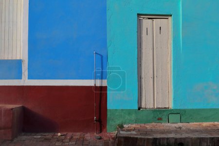 Foto de Trinidad, Cuba-12 de octubre de 2019: Fachada renovada pero rota de casa de estilo colonial cerca de la Plaza Mayor con una pequeña puerta de madera pintada de blanco y paredes coloreadas en combinaciones llamativas. - Imagen libre de derechos