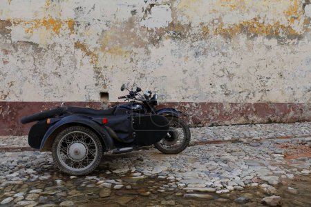 Photo for Trinidad, Cuba-October 13, 2019: Black MT-10-36 Dnepr sidecar motorcycle stationed on the cobbled, puddled Calle Alameda Street next to a chipped, paint-faded cream colored wall in the late afternoon. - Royalty Free Image
