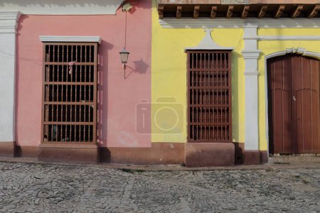 Trinidad, Cuba-October 13, 2019: Refurbished yet already chipped facades of colonial houses on Calle Amargura Street painted yellow and pink featuring brown wood door and grilles with turned balusters
