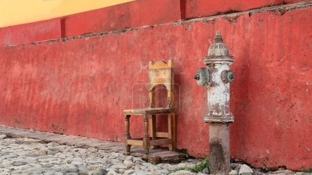 Trinidad, Cuba-October 13, 2019: Paint-faded, run down fire hydrant and old wooden chair on the narrow sidewalk of Calle Amargura Street south side, next to the SE corner with Calle Desengano Street.