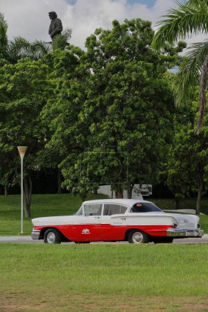 Photo for Santa Clara, Cuba-October 14, 2019: Old white with red American classic car -Chevrolet Delray 4 door Sedan from 1958- stationed on Plaza Ernesto Che Guevara Square, his statue jutting from the trees. - Royalty Free Image