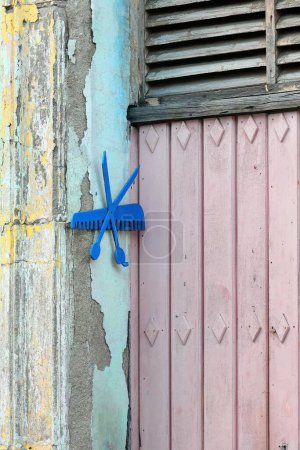 Camaguey, Kuba - 15. Oktober 2019: Friseursymbole - Kamm, Schere - werden neben einer rosa bemalten, geschlossenen Holztür eines Friseursalons im denkmalgeschützten historischen Zentrum an die abgesplitterte Wand genagelt..