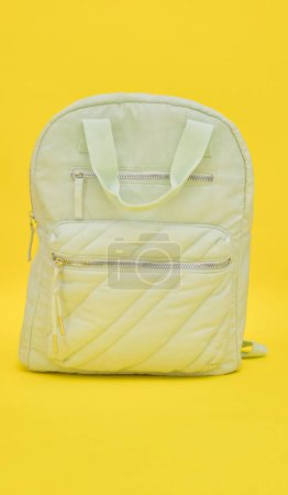 a light green backpack placed on a bright yellow background creates a striking contrast. The backpack is the center of attention, standing out against the bright yellow surface