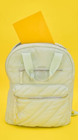 Una mochila blanca tendida en el suelo con una carpeta amarilla colocada encima de ella. La mochila y la carpeta son los principales temas de la imagen