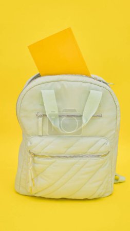 A white backpack lying on the ground with a yellow folder placed on top of it. The backpack and folder are the main subjects in the image