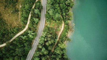 The aerial view shows a winding road snaking through the rugged mountains, surrounded by lush greenery and rocky terrain. The road twists and turns, offering a challenging drive through the scenic