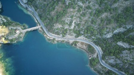The aerial view shows a winding road snaking through the rugged mountains, surrounded by lush greenery and rocky terrain. The road twists and turns, offering a challenging drive through the scenic