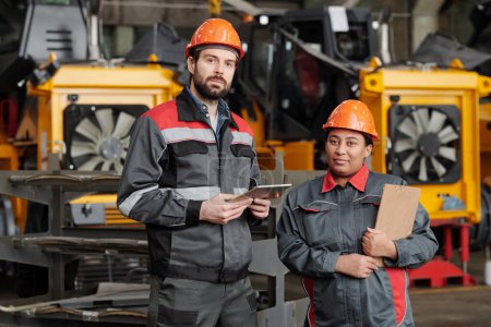 Foto de Two young interracial workers of contemporary plant or manufactory in hardhats and workwear standing by huge industrial machine - Imagen libre de derechos