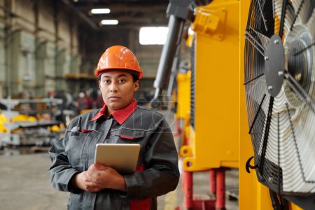 Foto de Young serious female worker of factory in hardhat and workwear holding digital tablet while checking industrial machines - Imagen libre de derechos