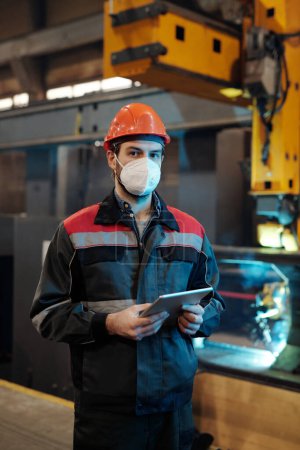 Foto de Young serious engineer in respirator, hardhat and workwear searching online technical data while using digital tablet by workplace - Imagen libre de derechos