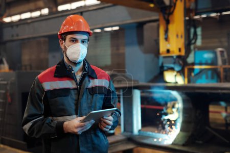 Photo for Contemporary factory engineer in protective workwear holding digital tablet while networking by his workplace in machinery plant - Royalty Free Image