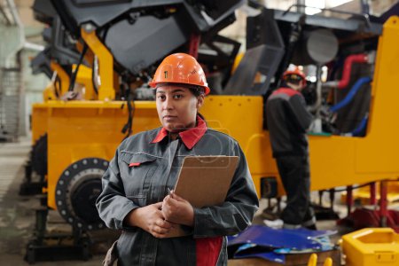 Téléchargez les photos : Confident African American female engineer with document standing against industrial machine in workshop and looking at camera - en image libre de droit