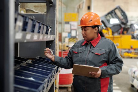 Foto de Serious young workwoman in safety helmet and uniform making revision of spare parts for industrial machines in modern plant - Imagen libre de derechos