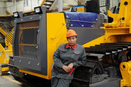 Foto de Serious young African American female worker in hardhat and workwear standing by caterpillar of huge industrial machine - Imagen libre de derechos
