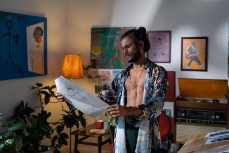Foto de Young inspirated man in non-gendered clothing looking at drawing while standing in studio of modern arts with paintings on walls - Imagen libre de derechos