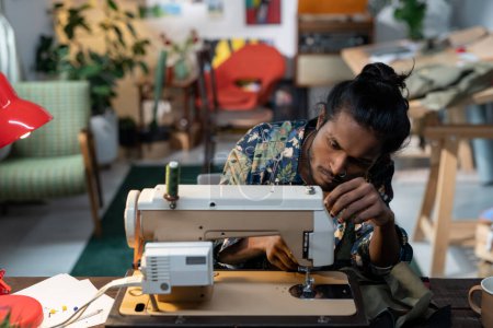 Foto de Young creative male tailor sitting by workplace in front of sewing machine while putting thread through needle eyes - Imagen libre de derechos