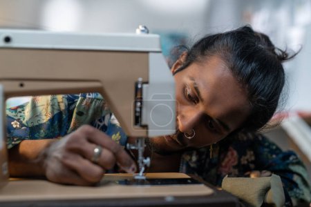 Foto de Serious guy of Indian ethnicity putting thread into needle eye of electric sewing machine while working over new fashion collection - Imagen libre de derechos