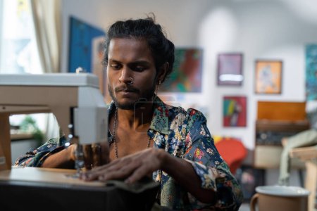 Foto de Serious stylish male tailor in non-gendered clothing creating new apparel while sitting in front of camera by electric sewing machine - Imagen libre de derechos