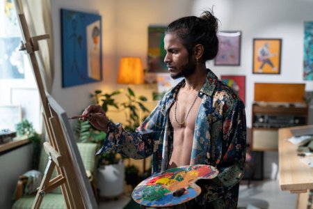 Téléchargez les photos : Young contemporary painter with color palette creating new artwork while standing in front of easel in his studio or workshop - en image libre de droit