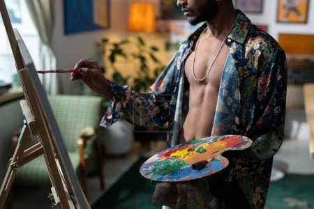 Foto de Middle section of young sexy man with color palette creating new painting on canvas while standing in front of easel - Imagen libre de derechos