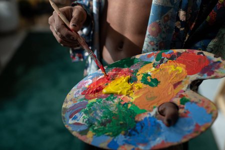 Téléchargez les photos : Hands of young non-binary man with paintbrush mixing acrylic paints on palette while creating new masterpiece in art studio - en image libre de droit