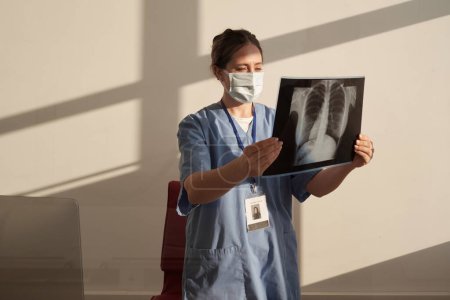 Téléchargez les photos : Young serious female radiologist looking at x-ray image after radiological examination of patient while standing in medical office - en image libre de droit