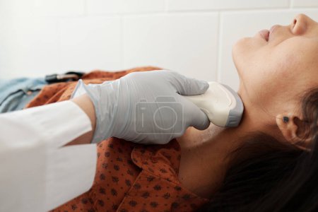 Foto de Gloved hand of doctor holding ultrasound scan on neck of young female patient during medical examination of thyroid in clinics - Imagen libre de derechos