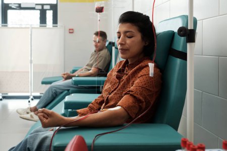 Foto de Young female and mature male donors giving blood for sick patients of modern hospital while sitting in armchairs in large ward - Imagen libre de derechos