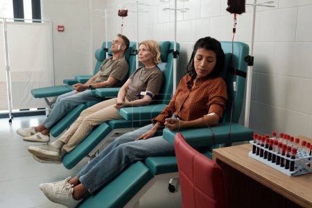 Foto de Group of blood donors with dropper tubes sitting in row on chairs in large hospital ward during procedure of hemotransfusion - Imagen libre de derechos