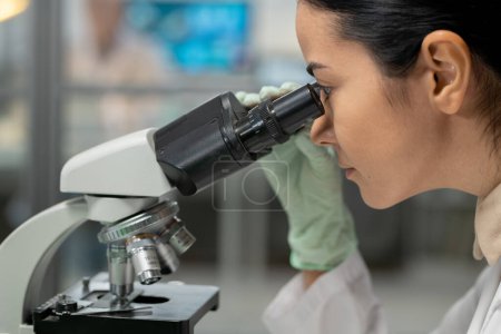 Photo for Young female scientist in protective gloves looking in microscope while studying new chemical substance and its characteristics - Royalty Free Image