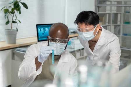 Photo for Two young interracial chemists in lab coats and protective masks looking at blue liquid in test tube while studying new compound - Royalty Free Image