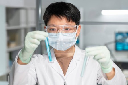 Photo for Asian female scientist in protective eyeglasses, mask and gloves comparing two samples of liquid substances - Royalty Free Image