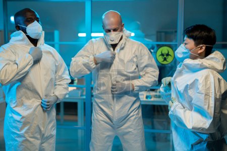 Photo for Group of interracial scientists putting on biohazard suits while standing in front of laboratory to work with dangerous substances - Royalty Free Image