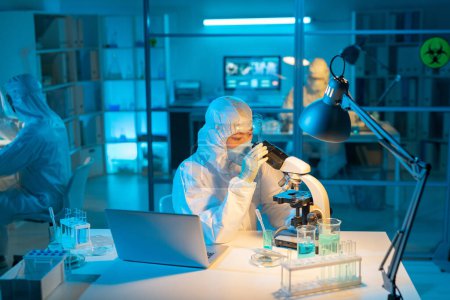 Photo for Female scientist in biohazard suit, respirator and gloves using microscope for studying new substances and their characteristics - Royalty Free Image