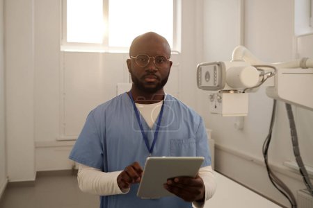 Téléchargez les photos : Young serious African American male clinician in uniform with tablet looking at camera while consulting patients online - en image libre de droit
