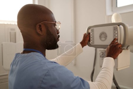 Téléchargez les photos : Young contemporary clinician in eyeglasses and blue uniform pressing button on control panel of medical equipment - en image libre de droit
