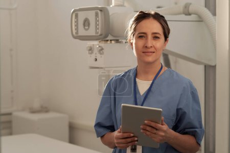 Téléchargez les photos : Happy young contemporary female clinician in blue uniform holding tablet while standing against medical equipment - en image libre de droit