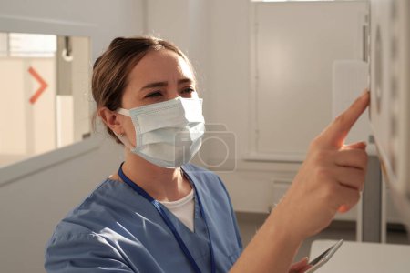 Foto de Young female nurse or radiologist in uniform and protective mask pushing button on panel of x-ray machine during work in clinics - Imagen libre de derechos