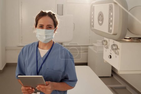 Téléchargez les photos : Young contemporary female radiologist in protective mask holding digital tablet while consulting clients online in hospital - en image libre de droit