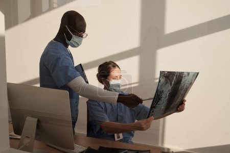 Téléchargez les photos : Young African American assistant in uniform and protective mask pointing at x-ray image held by female radiologist - en image libre de droit